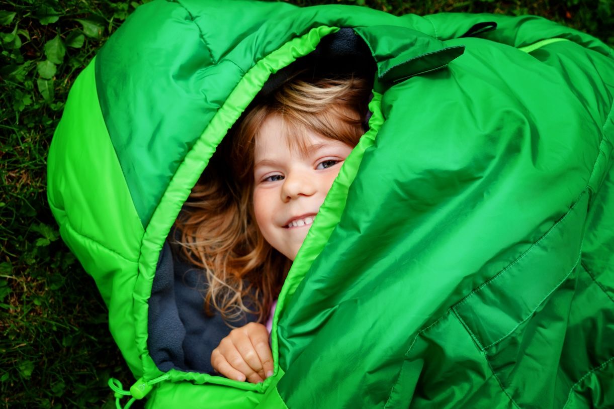 Child smiling inside a green sleeping bag outdoors, representing post-camping cleanup and sleeping bag care.