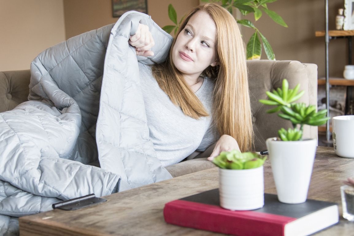 Young woman relaxing under a soft gray weighted blanket at home, showing comfort and care tips for washing weighted blankets.