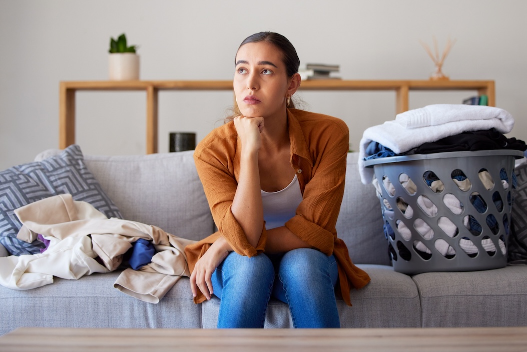 Tired woman sitting with overflowing laundry basket on couch at home, laundry is easier with Nashville laundry delivery services.